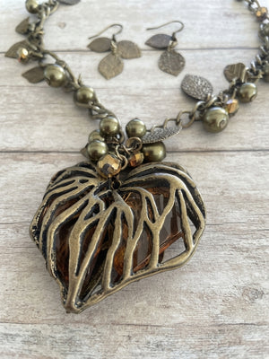 An antique bronze and gold-toned necklace with a large, organic leaf-shaped pendant featuring cutout branches, accompanied by matching dangling leaf earrings, displayed on a light wood background.