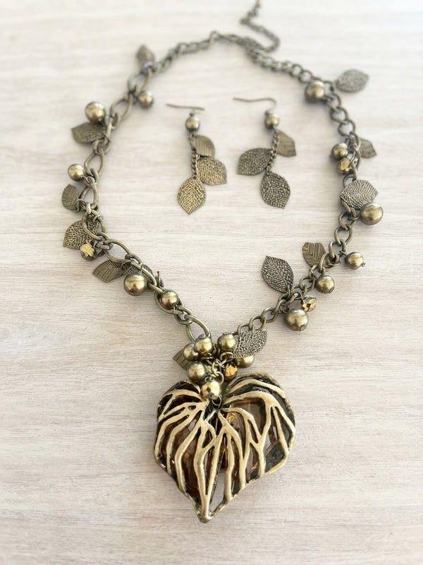 An antique bronze-toned necklace and matching drop earrings featuring textured leaf charms, small round beads, and a large leaf-shaped pendant with cutout details, displayed on a light wood surface.