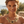 Charger l'image dans la galerie, A woman wearing a chunky gold statement necklace with floral details and black centers, photographed outdoors with a blurred coastal background.
