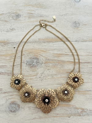 A gold-toned statement necklace featuring five ornate floral elements with dark centers and small clear rhinestones, displayed on a light wood background.