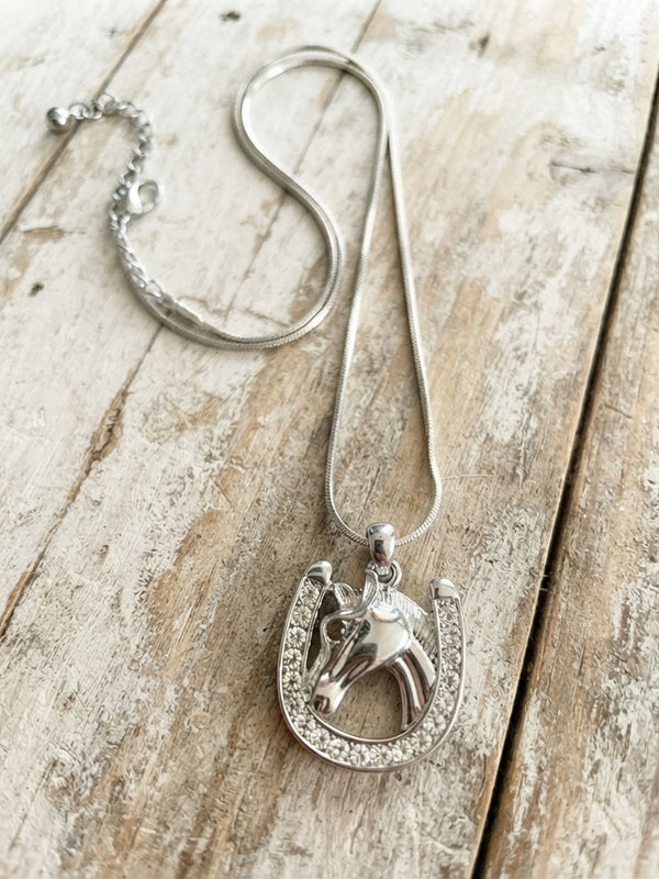A silver-toned necklace featuring a horse head charm set within a horseshoe-shaped outline, displayed on a rustic, light-colored wooden surface.