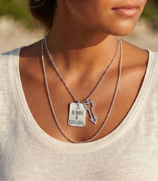 A close-up of a woman wearing a layered silver necklace set. The top layer is a delicate chain with a small silver key-shaped charm, while the bottom layer is a thicker chain with a rectangular dog tag pendant engraved with the words 'BE BRAVE & KEEP GOING'.