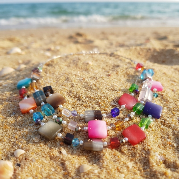 A colorful beaded necklace with square and round glass beads in pink, blue, green, purple, orange, and brown, displayed on a sandy beach with the ocean in the background.