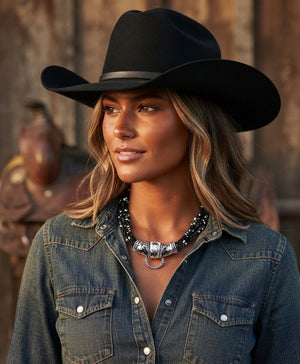 A woman wearing a black cowboy hat and a blue denim shirt, styled with a western silver necklace, set against a rustic wooden background.