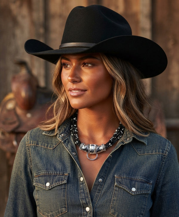 A woman wearing a black cowboy hat and a blue denim shirt, styled with a western silver necklace, set against a rustic wooden background.