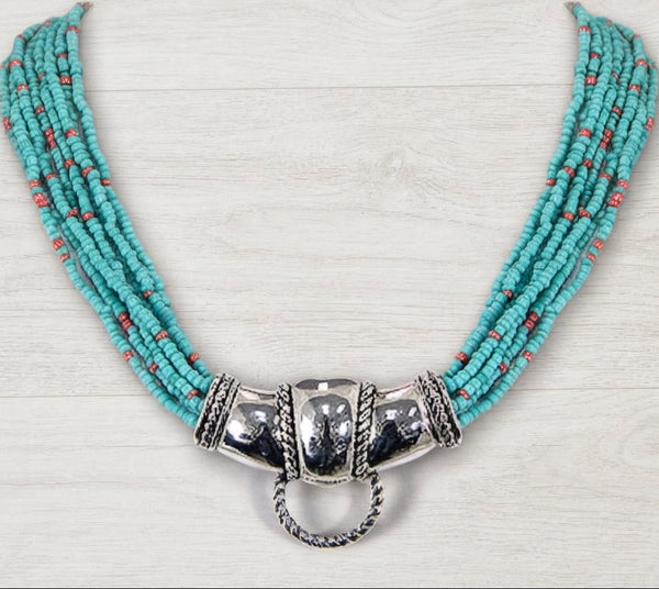 A multi-strand turquoise beaded necklace with a large polished silver-toned central clasp and hanging ring, displayed on a light wood background.