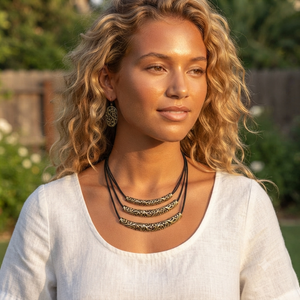 A woman with curly blonde hair wearing a multi-strand bohemian necklace with ornate gold-toned bars on a black cord, paired with matching dangling earrings and a white top, set against a soft-focus outdoor garden background.