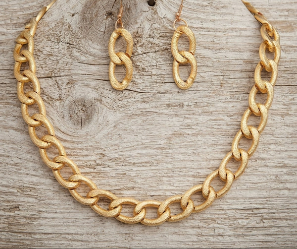A gold-tone chain necklace and matching drop earrings displayed on a weathered light wood surface, with a small nail behind the earrings as a styling prop.