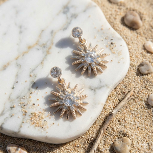 Gold sunburst earrings with clear stones on a marble surface with sand and pebbles.