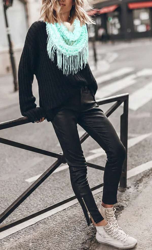 Person wearing a black sweater, green scarf, and black pants on a city street.