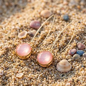 Pink earrings with gold accents on a sandy surface with seashells.