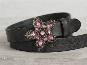 A black leather belt with a large, ornate silver-tone buckle featuring pink rhinestones, coiled on a light gray wooden surface.