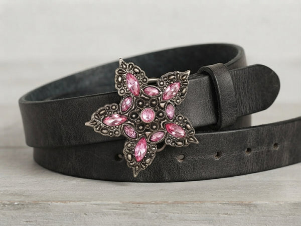 A black leather belt with a large, ornate silver-tone buckle featuring pink rhinestones, coiled on a light gray wooden surface.