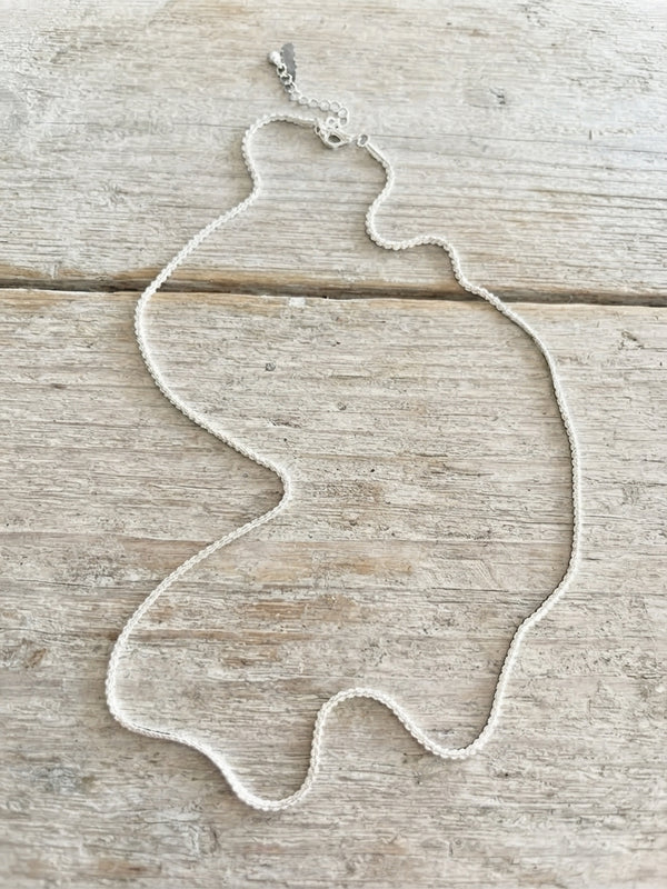 A delicate silver-toned beaded necklace with a lobster clasp, laid flat on a light-colored, rustic wooden surface.