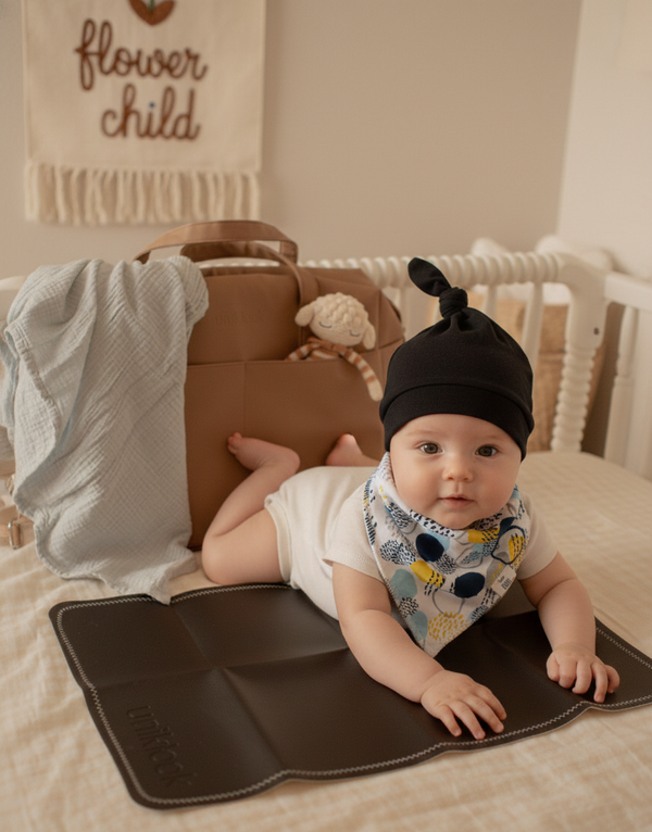 Baby lying on a changing mat with a black hat and colorful bib in a nursery setting.
