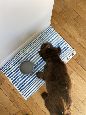 Dog standing on a blue and white striped rug with 'unikook' branding on a wooden floor.