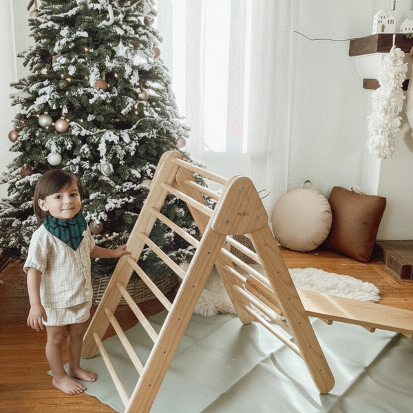 Child standing next to a wooden climbing toy in a room with a decorated Christmas tree.