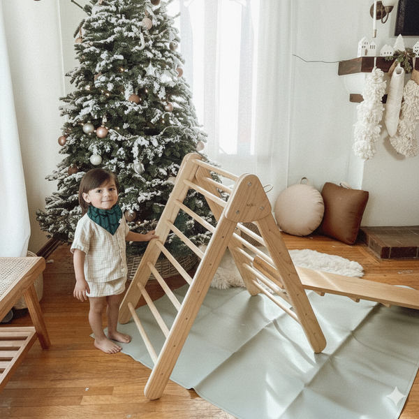 Child standing next to a wooden climbing toy in a room with a decorated Christmas tree.