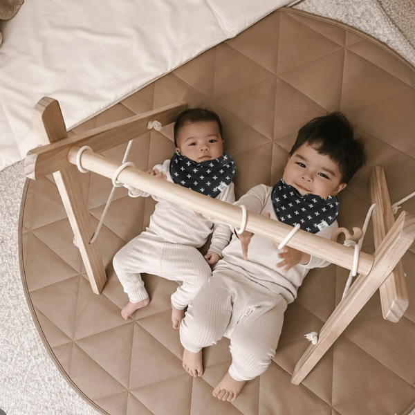 Two children lying on a wooden airplane toy inside a round, quilted playpen.