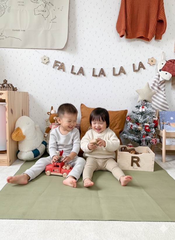Two children sitting on a green mat in a playroom with toys and Christmas decorations.