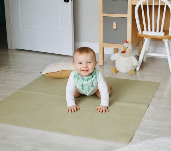 Baby crawling on a green mat in a room with furniture and toys.