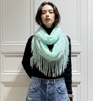 Person wearing a light blue scarf with white fringe in front of a white paneled wall.