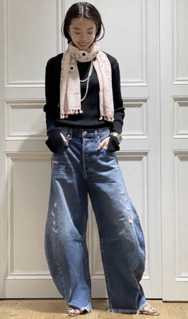 Person wearing a black sweater, blue jeans, and a pink scarf against a white paneled wall.