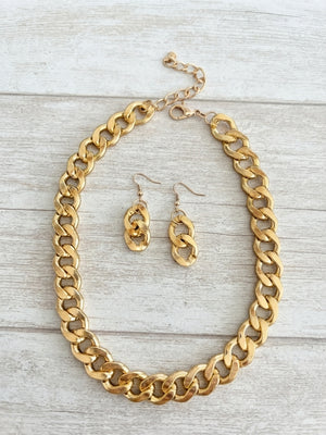A gold-tone chunky chain necklace and matching drop earrings displayed on a light gray wood plank background.