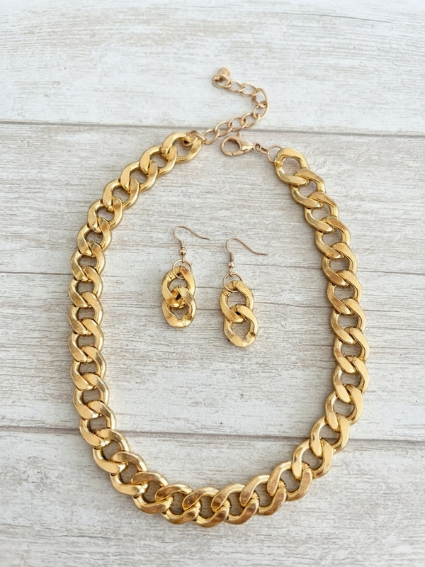 A gold-tone chunky chain necklace and matching drop earrings displayed on a light gray wood plank background.