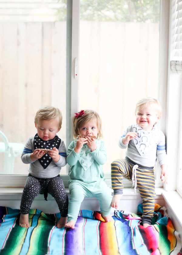 Three young children wearing matching striped bibs, sitting by a window and eating.
